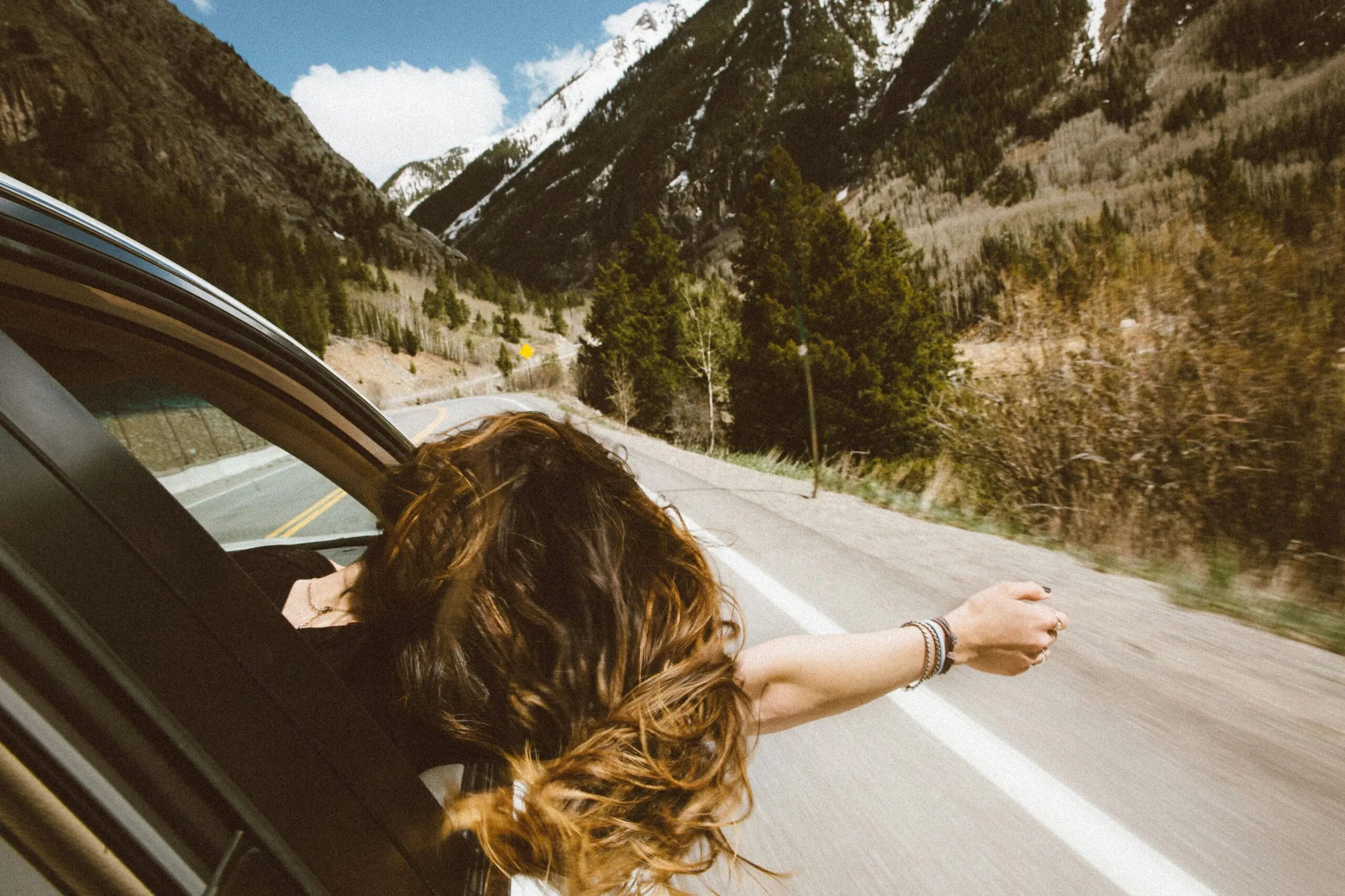 A person with long hair leans out of a car window, arm extended, on a scenic mountain road under a blue sky, conveying freedom and adventure.