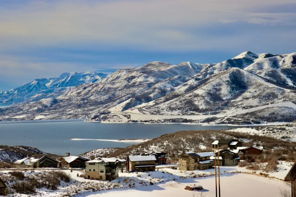 Snow-covered mountains and a calm lake create a serene winter scene, with several rustic houses nestled in the foreground under a cloudy sky.