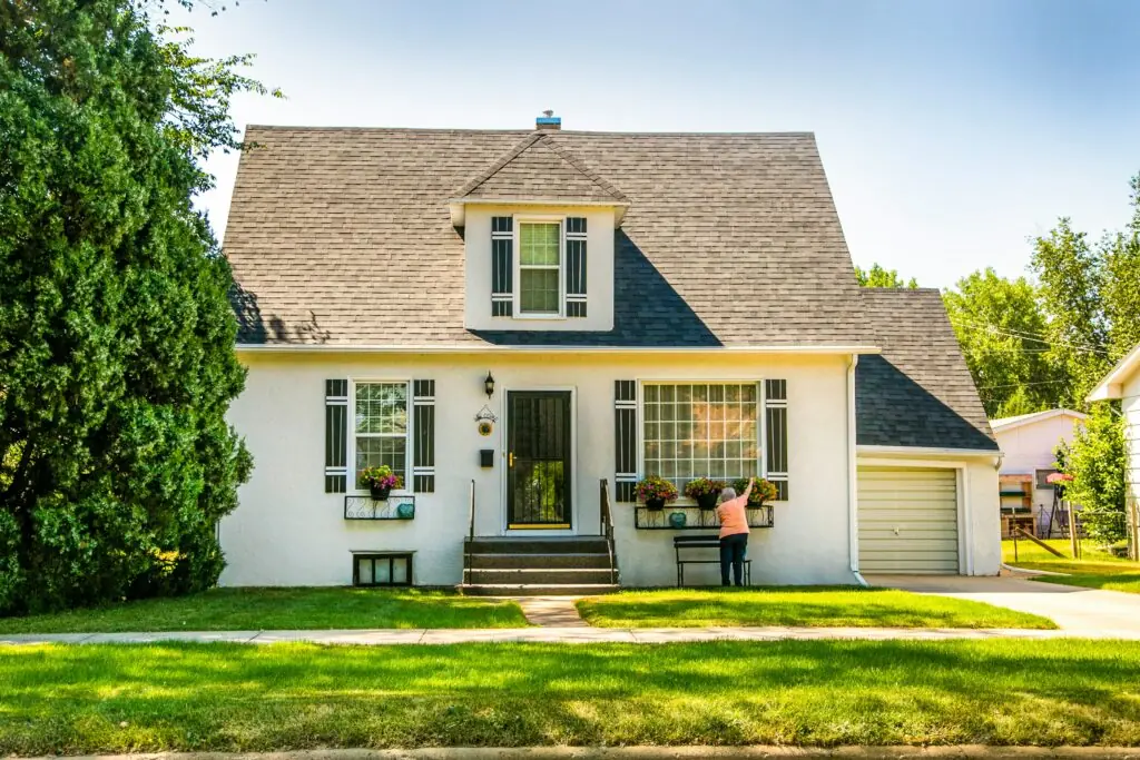 A charming, small white house with a steep grey roof. A person tends to flower boxes under large windows. Tall trees surround the house, creating a serene atmosphere.