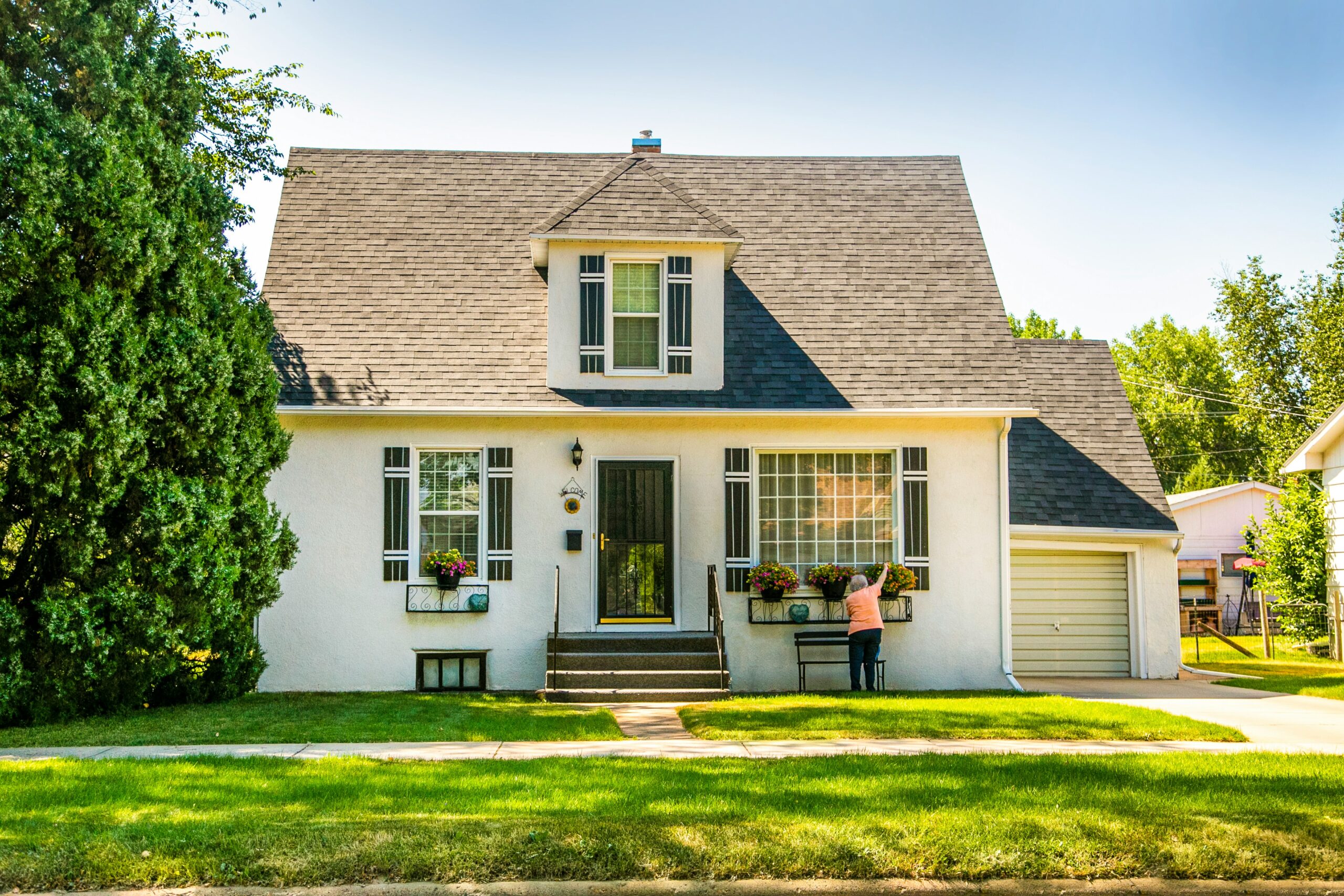 A charming, small white house with a steep grey roof. A person tends to flower boxes under large windows. Tall trees surround the house, creating a serene atmosphere.