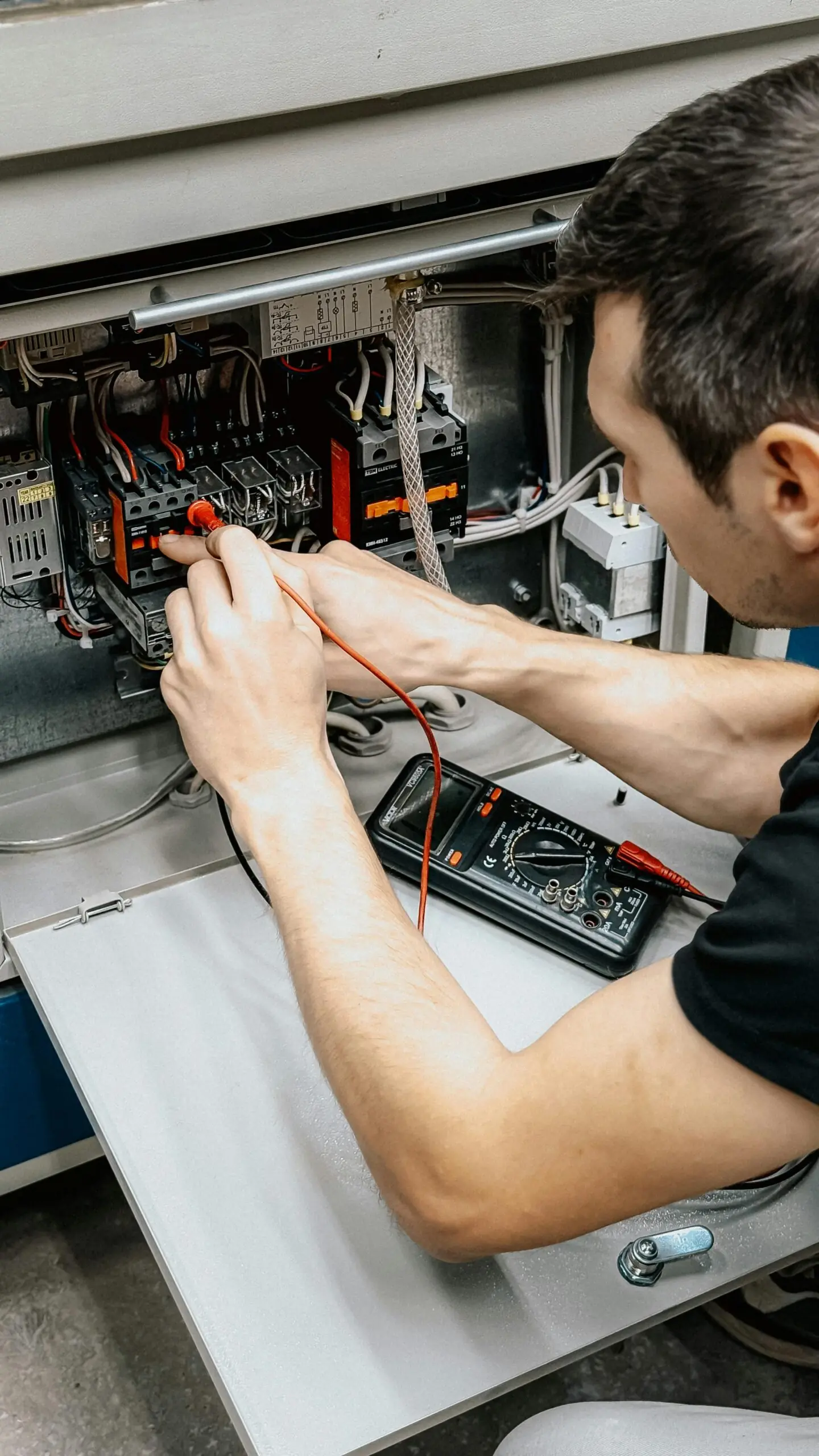 A technician uses a multimeter to test wires inside an electrical panel. They are focused, working in a workshop setting, conveying concentration and precision.
